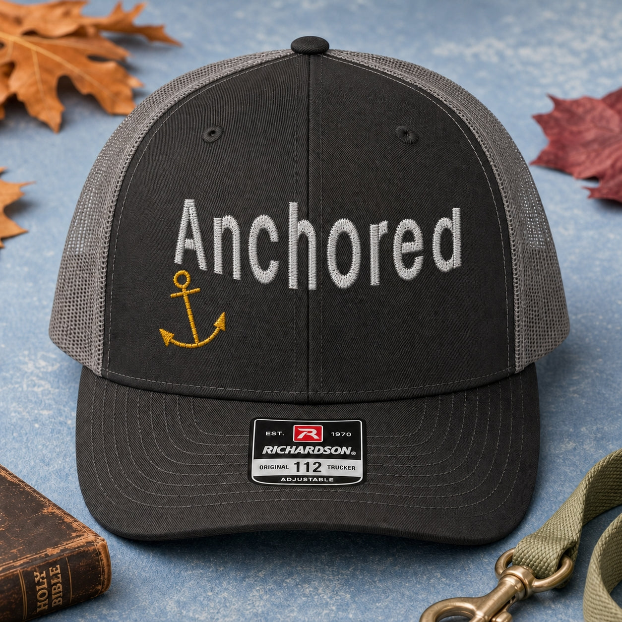 Anchored embroidery trucker hat in black and charcoal flatlay with Holy Bible, dog leash, and autumn leaves