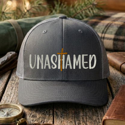 Gray cap with 'UNASHAMED' text on a wooden surface with outdoor items.