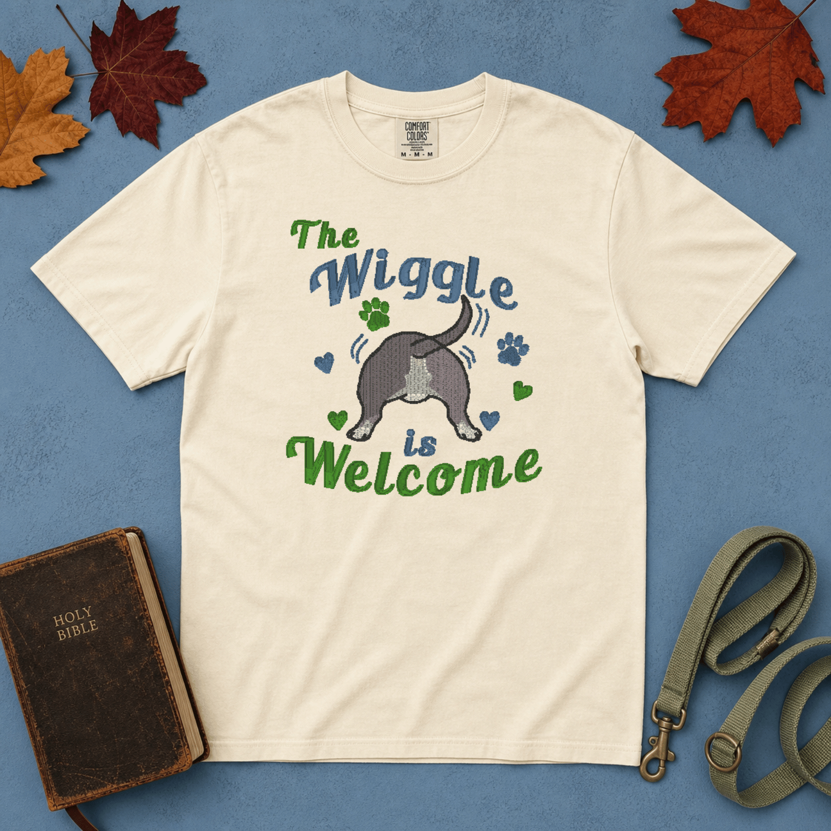 Ivory Comfort Colors tee with embroidered wiggle butt bully breed design, flatlay on blue background with Bible and leash
