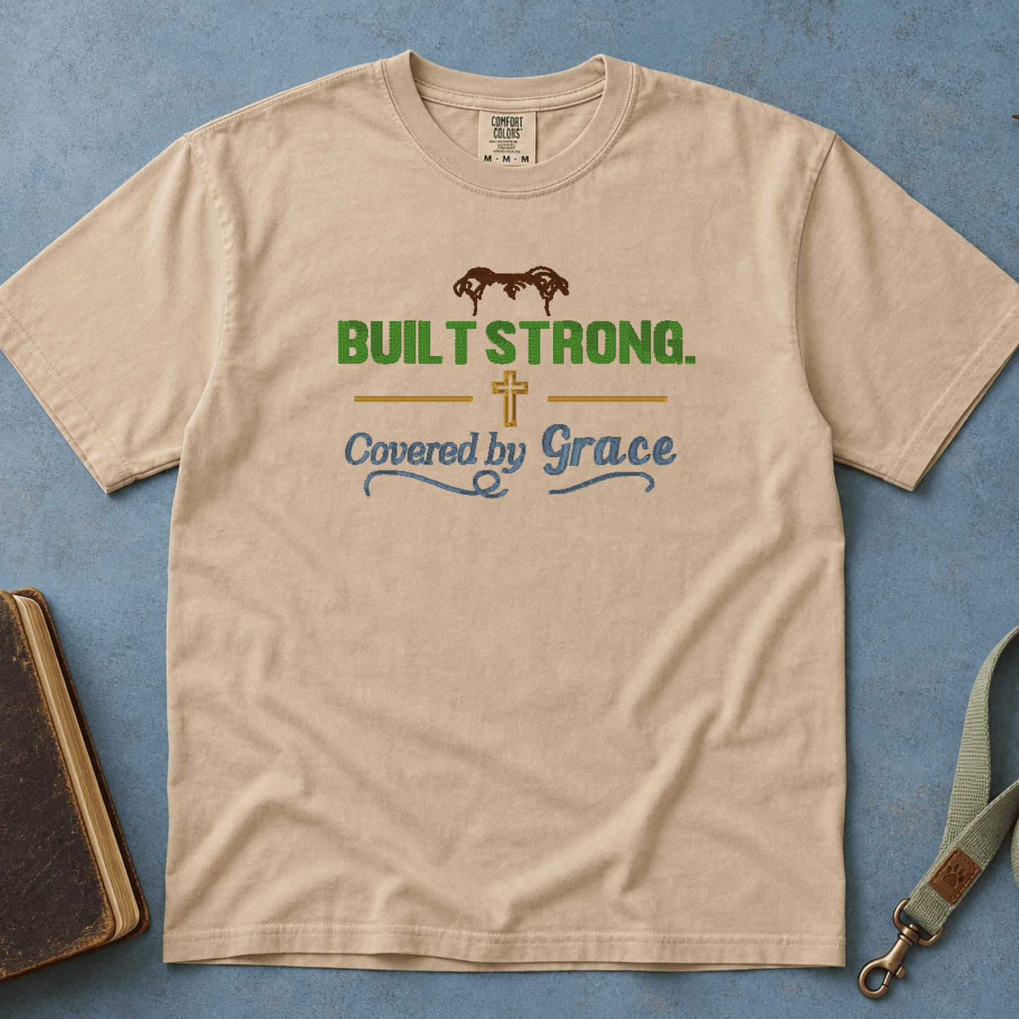 Built Strong Covered by Grace Comfort Colors Tee in Sandstone flatlay with Holy Bible and dog leash