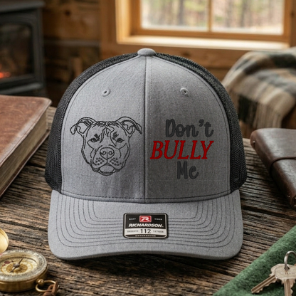 Gray cap with dog illustration and 'Don't Bully Me' text on a wooden surface with a cozy cabin background.