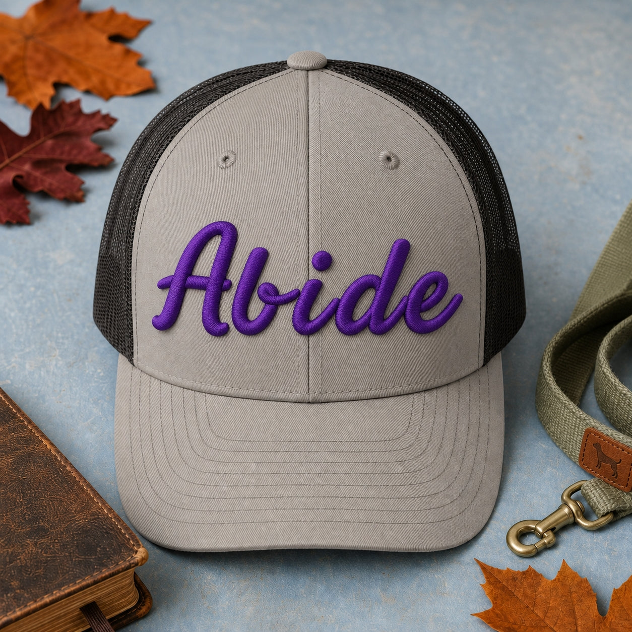 Abide puff embroidery trucker hat in grey and black flatlay with Holy Bible, dog leash, and autumn leaves