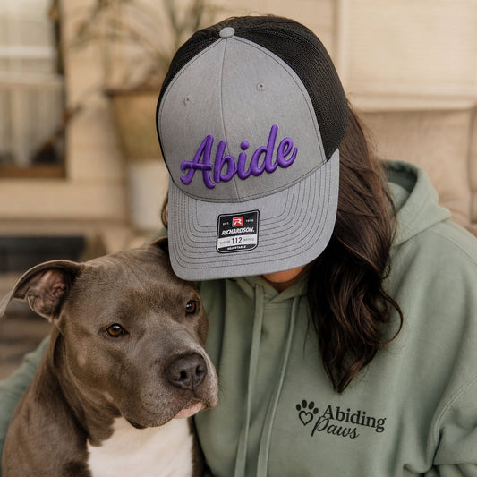 Woman wearing Abide puff embroidery grey and black Richardson 112 trucker hat with bully breed dog and Abiding Paws hoodie