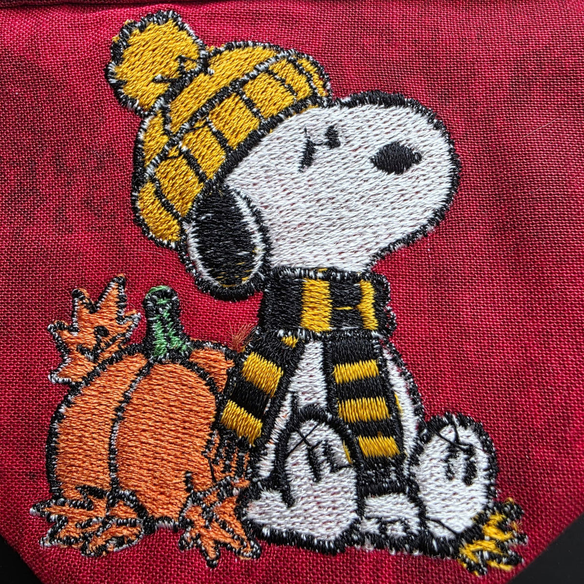 Close-up of embroidered white dog wearing golden knit hat and striped scarf beside pumpkin